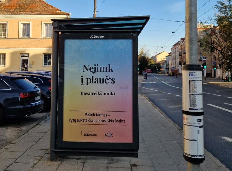 Nuotraukoje matomas viešojo transporto stotelės stendas su lauko reklama. Reklamos stende pateiktas užrašas „Nejimk į plaučius“. Skliaustuose pateikta posakio reikšmė – nesureikšmink. Stendo apačioje pateiktas prierašas: Pažink tarmes – rytų aukštaičių panevėžiškių šnekta.